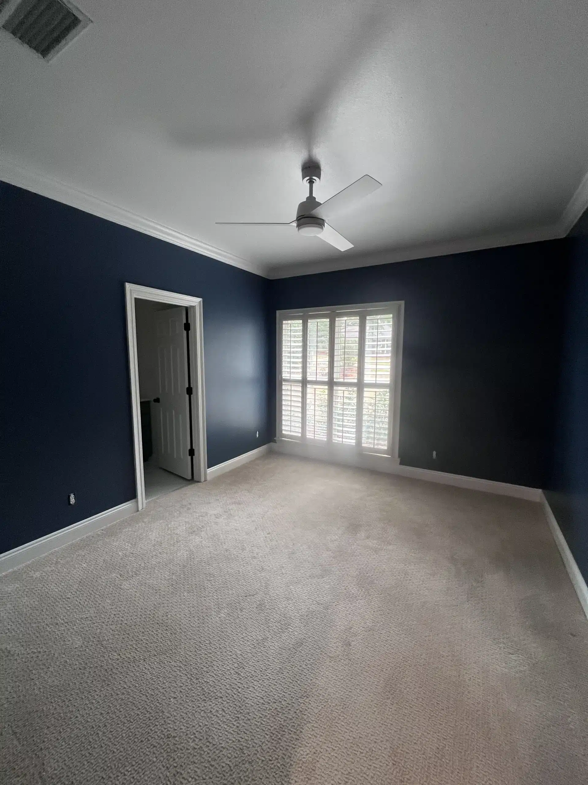 Professional Bedroom Painting with Dark Blue Walls and White Trim An empty bedroom showcasing dark blue walls, white trim, light carpet, and a ceiling fan after professional interior painting.